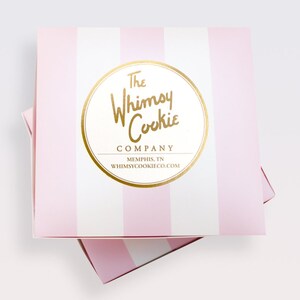 Puede incluir: Una caja de rayas rosas y blancas con un logotipo circular dorado que dice "The Whimsy Cookie Company, Memphis, TN, whimsycookieco.com"