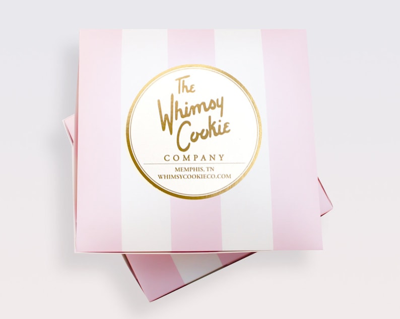 Puede incluir: Una caja de rayas rosas y blancas con un logotipo circular dorado que dice "The Whimsy Cookie Company, Memphis, TN, whimsycookieco.com"