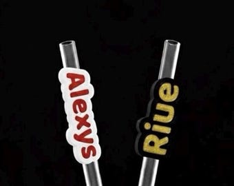 May include: Two clear drinking straws with personalized name tags. One tag reads "Alexys" in red letters on a white background. The other tag reads "Rive" in gold glitter letters on a black background. The straws are in a clear glass.
