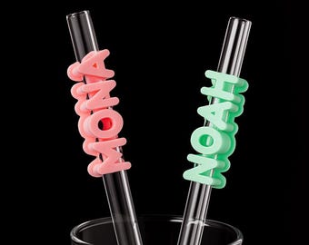 May include: Two clear glass straws with colorful silicone name tags. One straw has a pink tag that spells "MONA", and the other has a green tag that spells "NOAH". The straws are in a clear glass against a black background.
