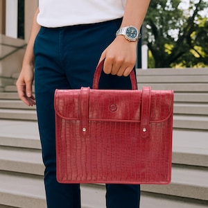 May include: A red briefcase with a crocodile skin texture is held by a person. The briefcase has a top handle and two straps with silver hardware. The person is wearing a white shirt, navy blue pants, and a silver watch.