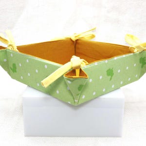 Puede incluir: Una cesta cuadrada de tela con exterior verde con lunares blancos y formas de dinosaurios verdes. El interior es naranja y cintas amarillas atadas en las esquinas. La cesta está sobre una superficie blanca.