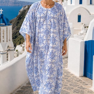 May include: A long, flowing caftan with a blue and white tile pattern. The caftan is worn by a person wearing sunglasses, walking on a stone path. The background features white buildings and a blue sea.
