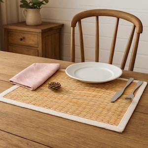 Könnte beinhalten: Ein gedeckter Tisch mit einem gewebten Tischset mit weißem Rand, einem weißen Teller, Besteck und einer rosa Serviette. Ein kleiner Tannenzapfen und ein Holzstuhl sind ebenfalls zu sehen. Der Tisch ist aus hellem Holz.