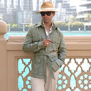 Puede incluir: Chaqueta safari verde claro con cinturón a juego, combinada con pantalones beige. La chaqueta tiene múltiples bolsillos y cierres de botón. Un sombrero de paja con una banda clara y gafas de sol oscuras completan el look. La persona sostiene un cigarro.