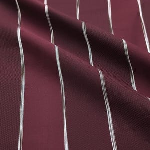 May include: A deep burgundy fabric featuring a textured weave and vertical pinstripes in a metallic silver tone. The material is draped to show folds and shadows, highlighting the contrast between the dark matte base and the reflective lines.