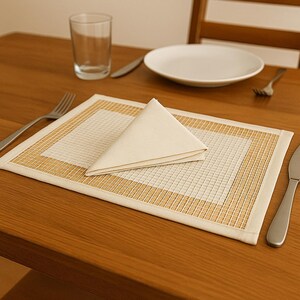 May include: A dining table setting with a folded cream napkin, a white plate, a glass, and a placemat with a woven border. A fork and knife are also present, creating a simple, elegant arrangement.