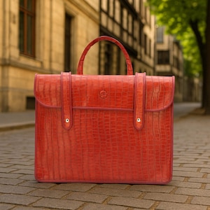 May include: A red briefcase with a textured, crocodile-like pattern. The bag has a top handle and a flap closure with a small logo. The bag has a rectangular shape and is standing on a brick surface.