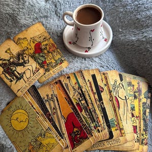 May include: A white ceramic coffee cup with a small heart pattern on a matching saucer, positioned above a spread of vintage-style tarot cards. The cards feature detailed illustrations, including the Sun, and text such as Kupa Ş&ouml;valyesi and Ay.