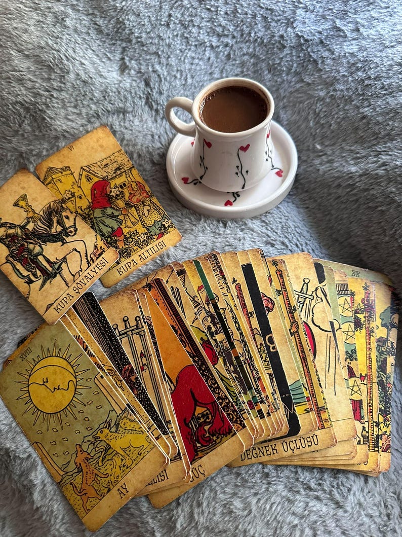 May include: A white ceramic coffee cup with a small heart pattern on a matching saucer, positioned above a spread of vintage-style tarot cards. The cards feature detailed illustrations, including the Sun, and text such as Kupa Ş&ouml;valyesi and Ay.