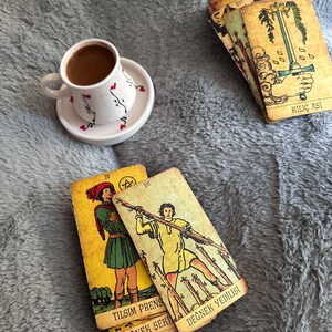 May include: A white ceramic cup and saucer with small red heart and black vine patterns containing dark coffee, alongside several tarot cards with yellow backgrounds and illustrations, including cards labelled KILI&Ccedil; ASI, TILSIM PRENS, and DEĞNEK YEDİLİSİ.