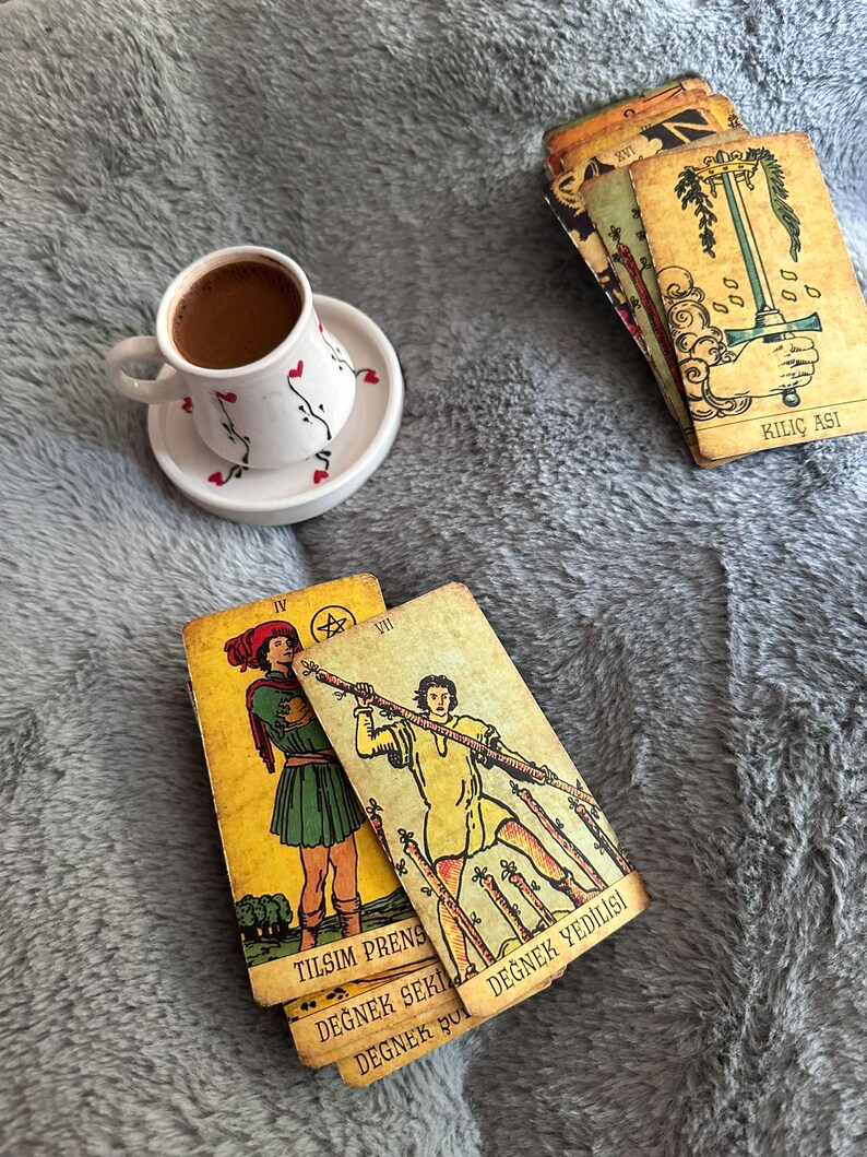 May include: A white ceramic cup and saucer with small red heart and black vine patterns containing dark coffee, alongside several tarot cards with yellow backgrounds and illustrations, including cards labelled KILI&Ccedil; ASI, TILSIM PRENS, and DEĞNEK YEDİLİSİ.