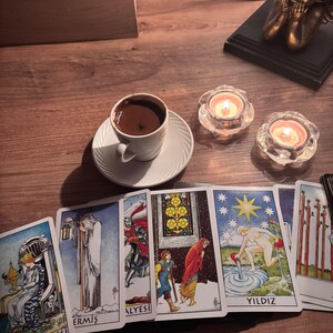 May include: A white ceramic cup filled with dark coffee on a matching saucer, positioned next to two lit tea light candles in glass holders and a row of six tarot cards featuring colourful illustrations and the text Kupa Krali&ccedil;esi, Ermiş, and Yildiz.