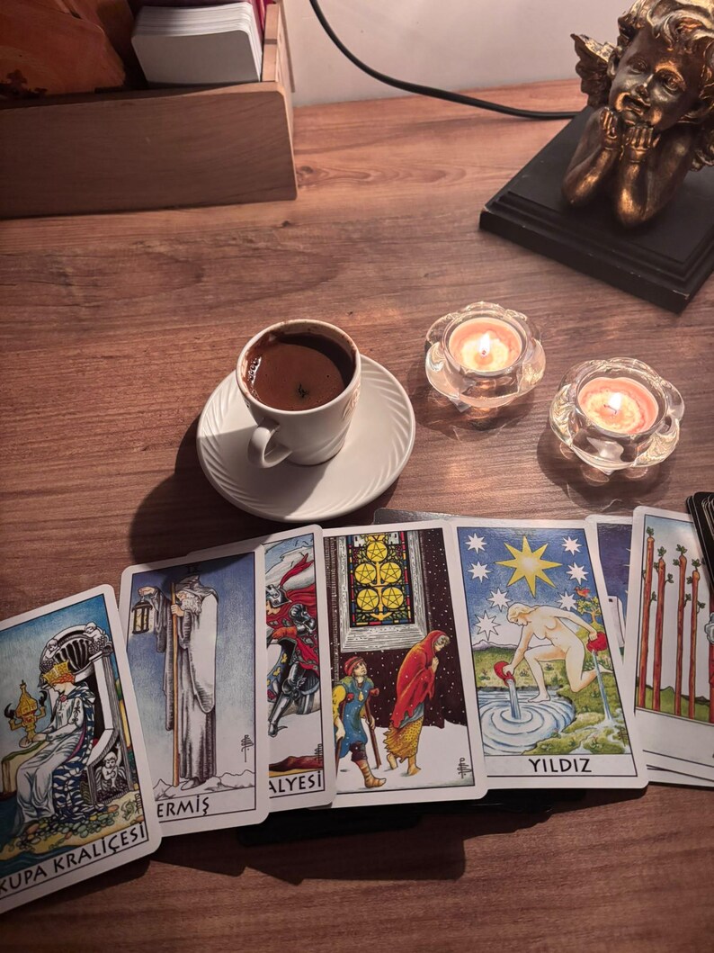 May include: A white ceramic cup filled with dark coffee on a matching saucer, positioned next to two lit tea light candles in glass holders and a row of six tarot cards featuring colourful illustrations and the text Kupa Krali&ccedil;esi, Ermiş, and Yildiz.