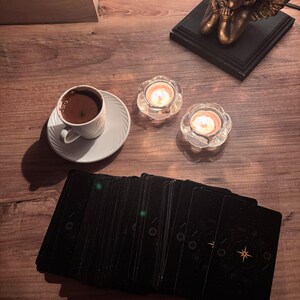 May include: A deck of black tarot cards with gold celestial designs, a white ceramic cup filled with dark coffee on a matching saucer, two lit tea light candles in clear glass holders, and a bronze-coloured cherub figurine on a black base.