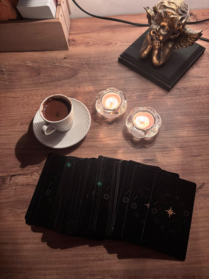 May include: A deck of black tarot cards with gold celestial designs, a white ceramic cup filled with dark coffee on a matching saucer, two lit tea light candles in clear glass holders, and a bronze-coloured cherub figurine on a black base.