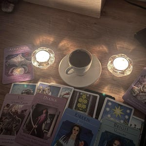 May include: A white ceramic cup filled with dark coffee sits on a matching saucer between two lit glass tea light holders. Several oracle cards with illustrations and text, including names like Aeracura, Isis, Diana, Eireen, and Brigit, are spread out.