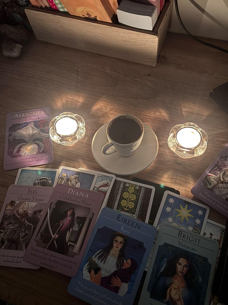 May include: A white ceramic cup filled with dark coffee sits on a matching saucer between two lit glass tea light holders. Several oracle cards with illustrations and text, including names like Aeracura, Isis, Diana, Eireen, and Brigit, are spread out.