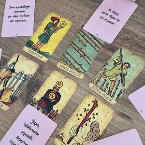 May include: A collection of tarot cards and rectangular pink message cards with Turkish text arranged on a wooden surface. The tarot cards include the Page of Pentacles, Eight of Wands, Seven of Wands, Four of Pentacles, and Ace of Wands.