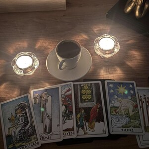 May include: A white ceramic coffee cup on a saucer, two lit tea light candles in glass holders, and a spread of tarot cards including Kupa Krali&ccedil;esi, Ermiş, and Yildiz. The cards feature detailed illustrations and text labels on a wooden surface.