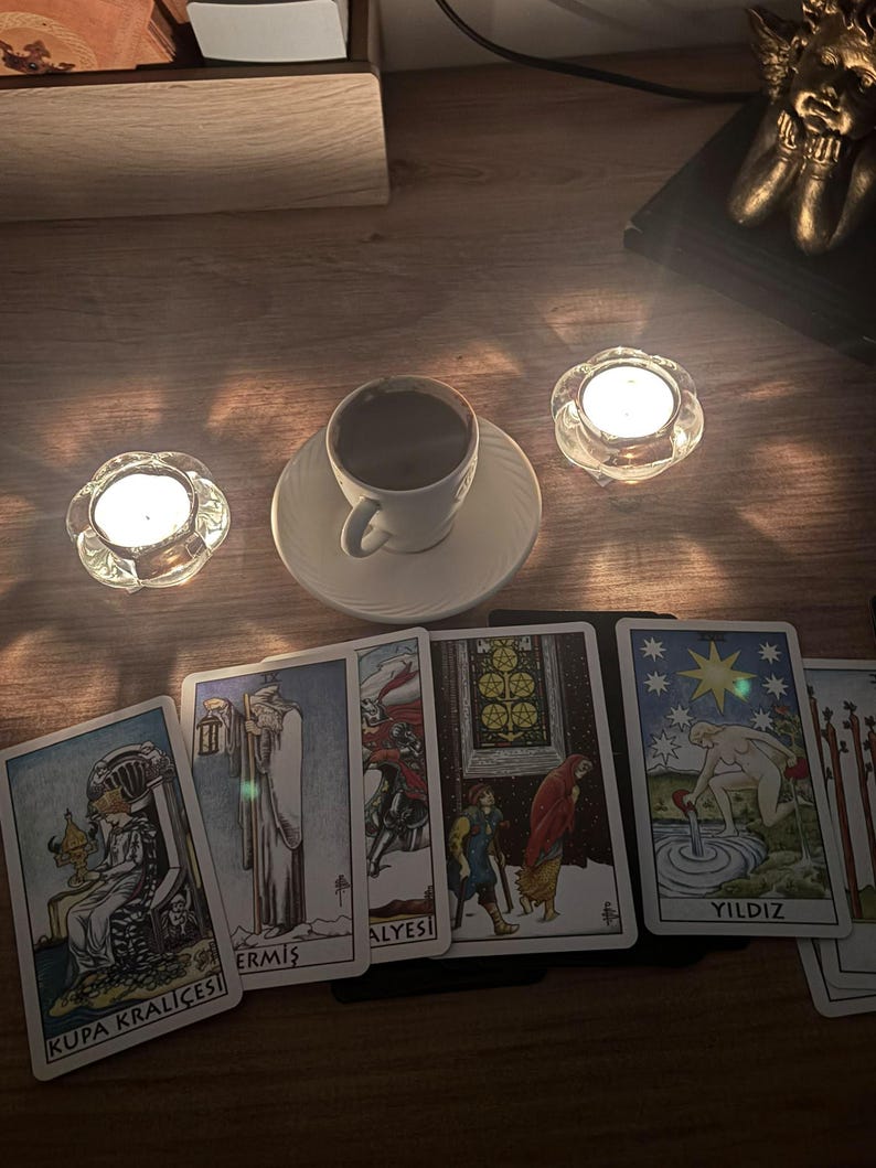May include: A white ceramic coffee cup on a saucer, two lit tea light candles in glass holders, and a spread of tarot cards including Kupa Krali&ccedil;esi, Ermiş, and Yildiz. The cards feature detailed illustrations and text labels on a wooden surface.