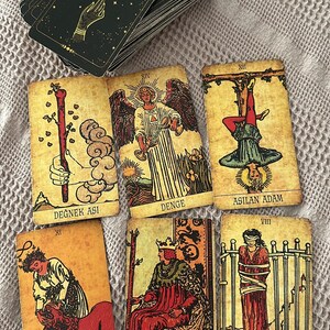 May include: A deck of tarot cards with black backs featuring a gold hand and sun design, alongside six face-up cards labelled Degnek Asi, Denge, Asilan Adam, Guc, Degnek Krali, and Kilic Sekizlisi, each displaying detailed vintage-style illustrations.