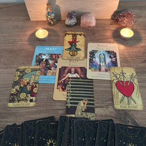 May include: A tarot card spread featuring multiple cards including Maat, The Hanged Man, Bast, Isolt, Ten of Pentacles, Nine of Swords, and Three of Swords. Two lit tea light candles, crystals, and a wooden box containing card decks are present.