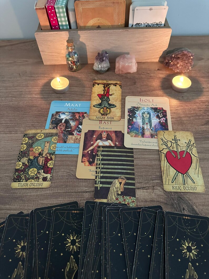 May include: A tarot card spread featuring multiple cards including Maat, The Hanged Man, Bast, Isolt, Ten of Pentacles, Nine of Swords, and Three of Swords. Two lit tea light candles, crystals, and a wooden box containing card decks are present.