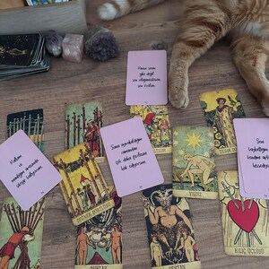 May include: A collection of tarot cards spread out on a wooden surface, including traditional illustrated cards like The Lovers, The Devil, and The Star, alongside several light pink cards featuring handwritten Turkish text messages.