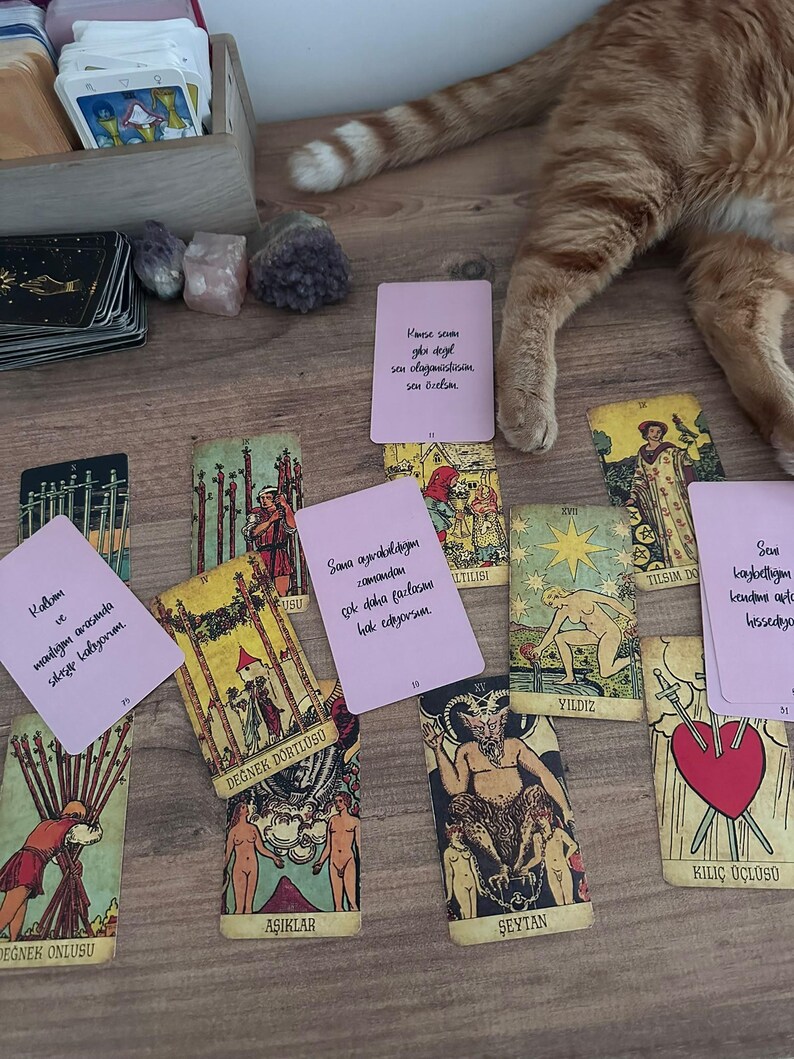 May include: A collection of tarot cards spread out on a wooden surface, including traditional illustrated cards like The Lovers, The Devil, and The Star, alongside several light pink cards featuring handwritten Turkish text messages.