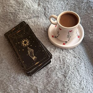 May include: A stack of black tarot cards featuring gold celestial sun and hand illustrations alongside a white ceramic cup and saucer decorated with small red hearts and black vine patterns, filled with dark brown coffee.