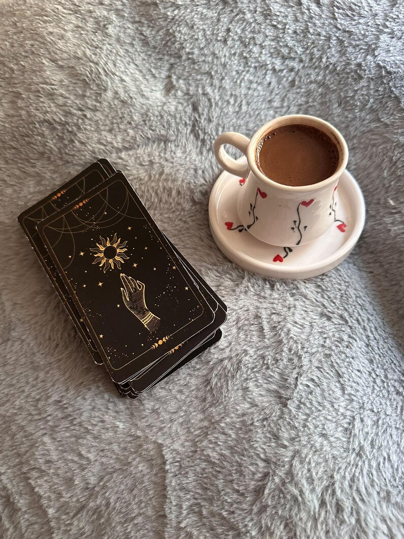 May include: A stack of black tarot cards featuring gold celestial sun and hand illustrations alongside a white ceramic cup and saucer decorated with small red hearts and black vine patterns, filled with dark brown coffee.