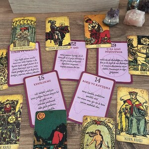 May include: A collection of tarot cards spread out on a surface, including several cards with Turkish text and illustrations. The cards feature various figures, symbols, and colours, with a wooden box filled with additional card decks positioned at the top.