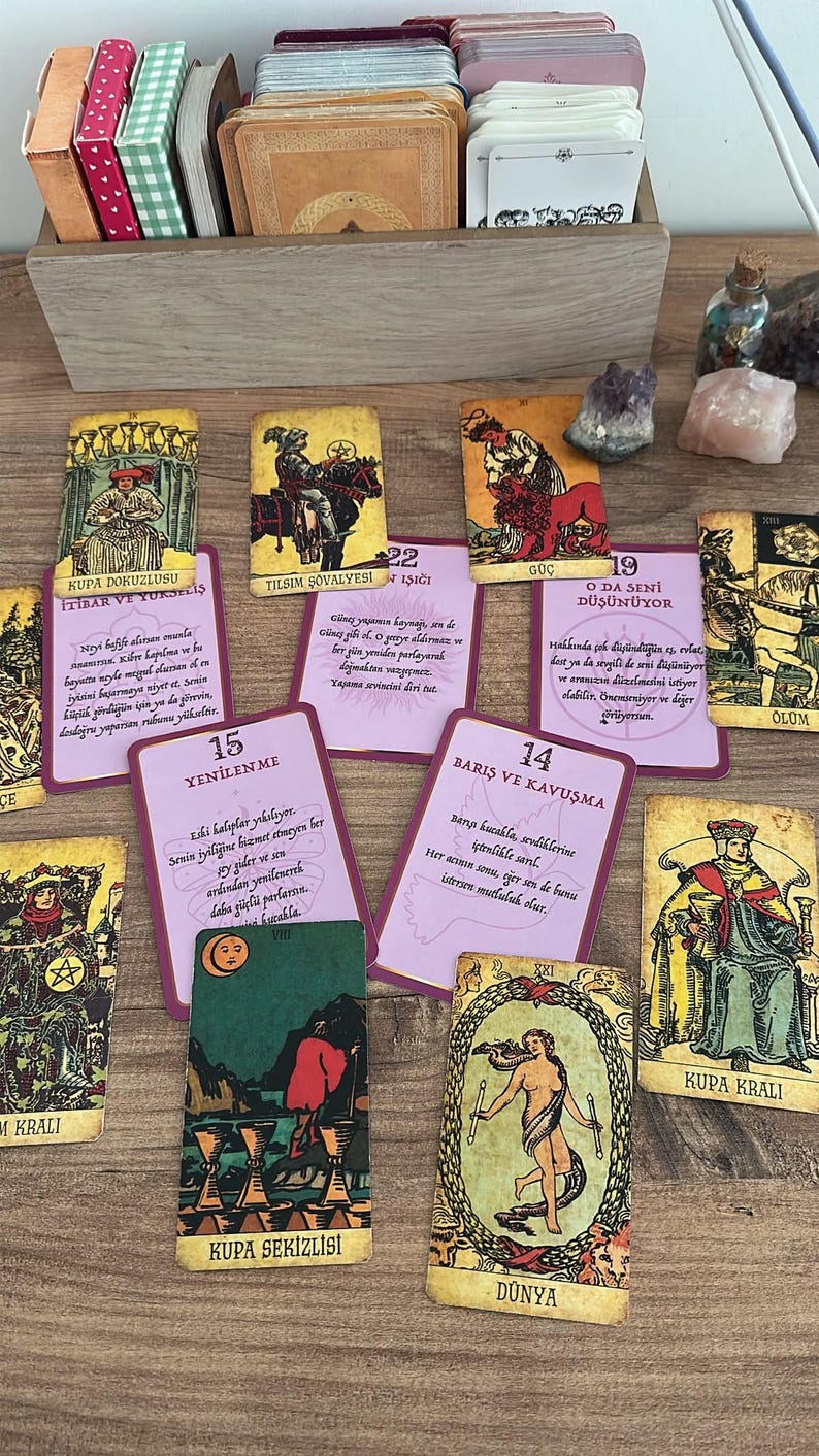 May include: A collection of tarot cards spread out on a surface, including several cards with Turkish text and illustrations. The cards feature various figures, symbols, and colours, with a wooden box filled with additional card decks positioned at the top.