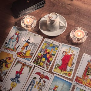 May include: A collection of tarot cards featuring illustrations including Kili&ccedil; Kralı, Asilan Adam, and Değnek Prensi, arranged alongside a white coffee cup on a saucer, a stack of black cards, and two lit candles in clear glass holders.
