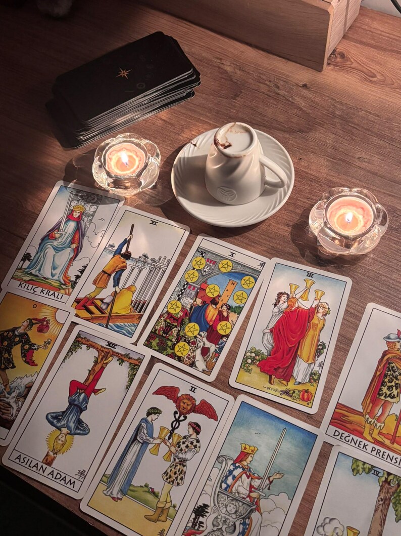 May include: A collection of tarot cards featuring illustrations including Kili&ccedil; Kralı, Asilan Adam, and Değnek Prensi, arranged alongside a white coffee cup on a saucer, a stack of black cards, and two lit candles in clear glass holders.