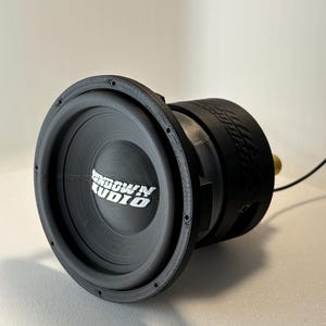May include: A black subwoofer with the text "Sundown Audio" in white. The speaker has a circular design with a black surround and a cylindrical body. A gold-colored connector is visible.