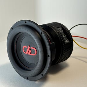 May include: A black subwoofer with a red logo and accents. The subwoofer has a circular design with a ridged surround and a series of small holes around the edge. Wires extend from the back.