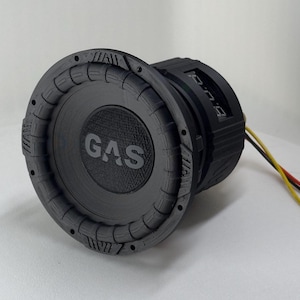 May include: A black circular subwoofer speaker featuring a textured cone with the word GAS printed in the center. The outer rim has a ridged design with small circular holes. A digital display is visible on the side, with yellow and black wires.