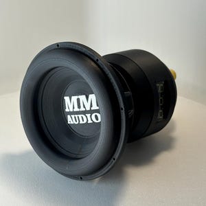 May include: A black subwoofer with the text "MM AUDIO" in white. The speaker has a circular design with a ribbed edge and a cylindrical body. A small yellow component is visible on the side.