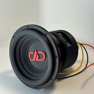 May include: A black subwoofer with a red "JD" logo on the speaker cone. The speaker has a circular design with multiple screw holes around the edge. Red, yellow, and black wires extend from the back.