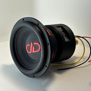 May include: A black subwoofer with a red accent ring and a red "DD" logo. The speaker has a circular design with a black frame and several screw holes. Wires in yellow, red, and black extend from the back of the speaker.