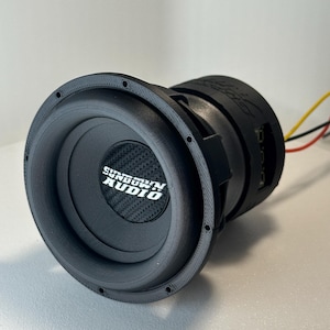 Op de afbeelding: Een zwarte Sundown Audio subwoofer met een koolstofvezel conus. De subwoofer heeft een cirkelvormig ontwerp met een getextureerde buitenrand en de merknaam in het wit gedrukt. Draden lopen van de achterkant.