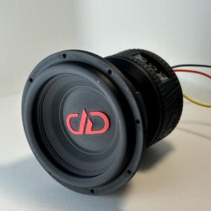 May include: A black subwoofer with a red logo on the speaker cone. The subwoofer has a circular design with a ribbed texture around the speaker. Wires extend from the back of the subwoofer.
