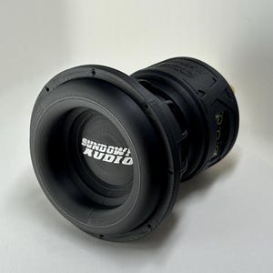 May include: A black Sundown Audio subwoofer with a circular design. The subwoofer has a large, round opening on one side and a smaller opening on the other. The Sundown Audio logo is visible in white text.