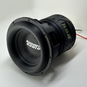 May include: A black Sundown Audio subwoofer with a textured design. The speaker has a circular shape with a central cone and a surrounding rim. The text "Sundown Audio" is visible on the speaker. Wires extend from the back.