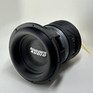 May include: A black Sundown Audio subwoofer with a circular design. The subwoofer has a prominent cone and a textured surround. The brand name is displayed in white lettering on the front. A yellow wire extends from the back.
