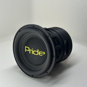 Op de afbeelding: Een zwarte subwoofer-luidspreker met een cirkelvormig frame en een gestructureerde koolstofvezel stofkap. Het woord Pride is in felgele letters gedrukt in het midden van de luidsprekerconus. De unit heeft een diepe cilindrische behuizing.