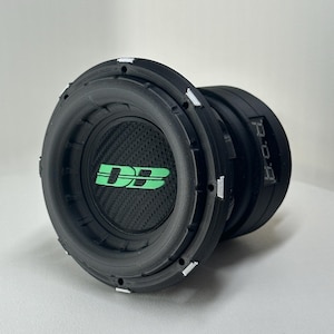 Pode incluir: Um subwoofer preto com um cone de fibra de carbono e um logotipo verde. O subwoofer tem um design circular com uma série de detalhes brancos na borda. O texto "D.B." é visível em verde na frente do alto-falante.