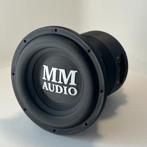 May include: A black subwoofer with the text "MM AUDIO" in white. The speaker has a circular design with a textured rim and a central cone. The speaker is designed for audio systems.